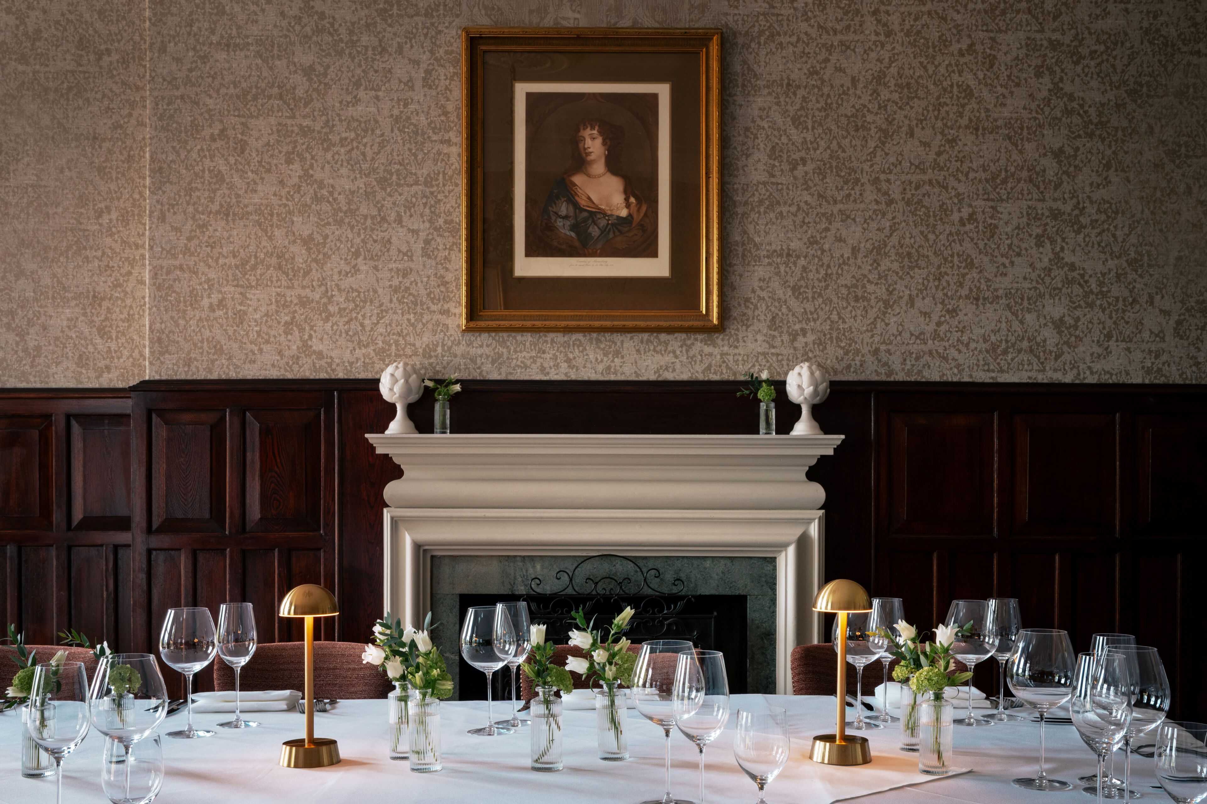 Une table à manger dressée avec des verres à vin, une lampe dorée et un portrait encadré accroché au mur.
