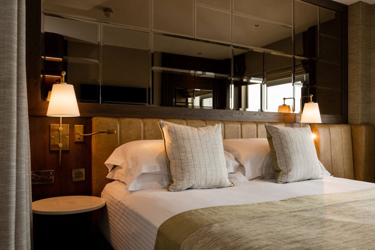 A hotel room with a large bed, a round side table, a mirror above the bed, and a wall-mounted lamp.