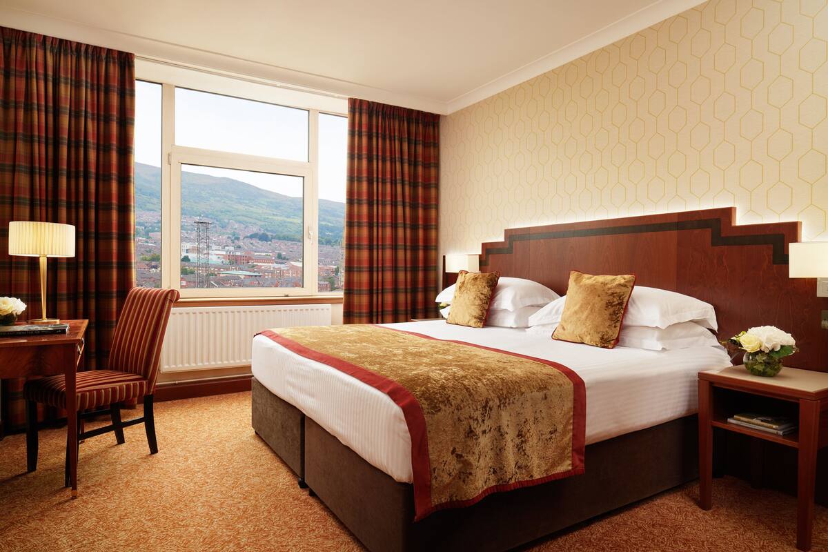 A hotel room with a large bed, a desk with a chair, a nightstand, and a view of a cityscape through a window.