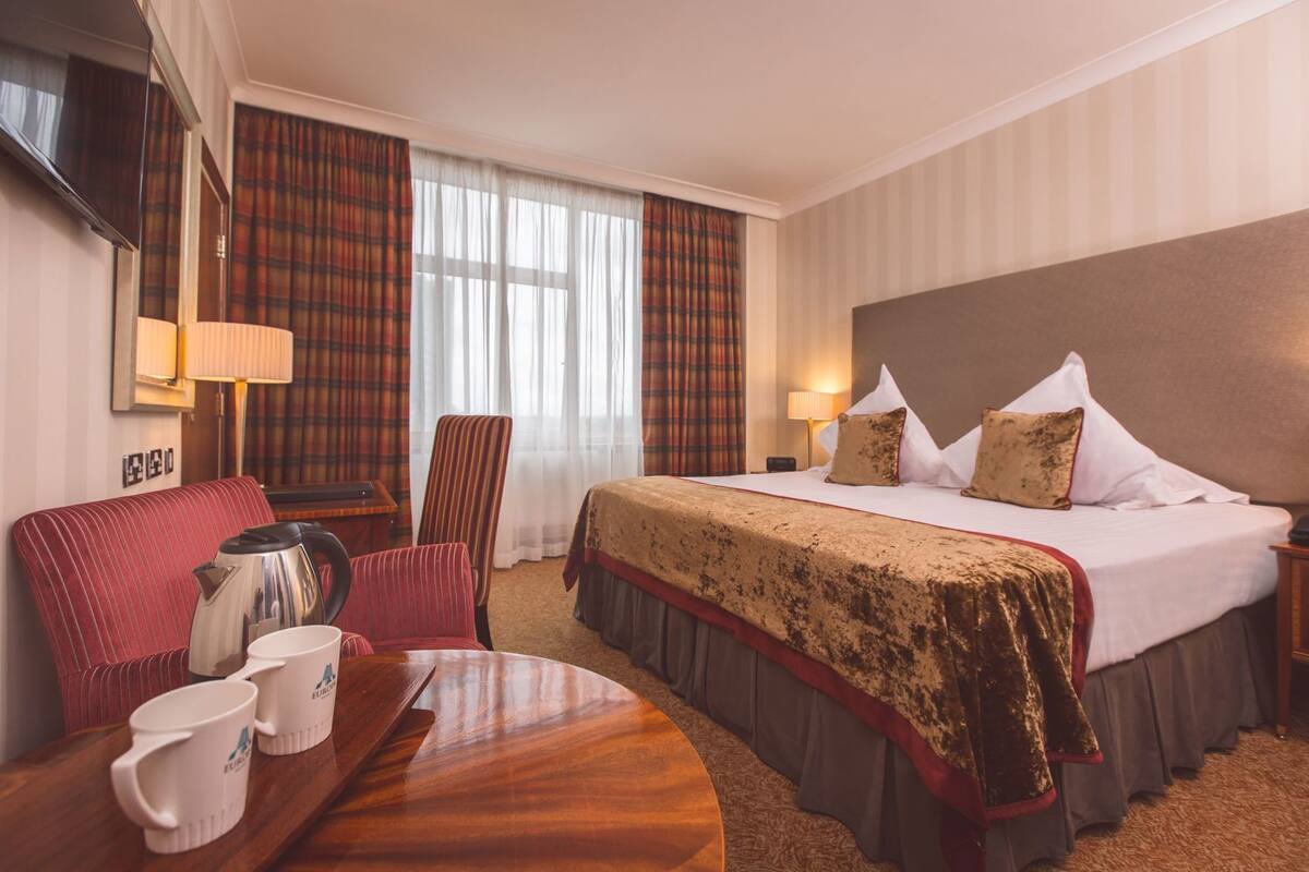 A hotel room with a bed, a desk with a kettle, a chair, and a table with mugs.
