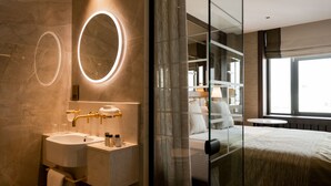A modern bathroom with a marble vanity, a large mirror, and a glass partition leading to a bedroom with a bed and a nightstand. - Europa Hotel (Belfast)