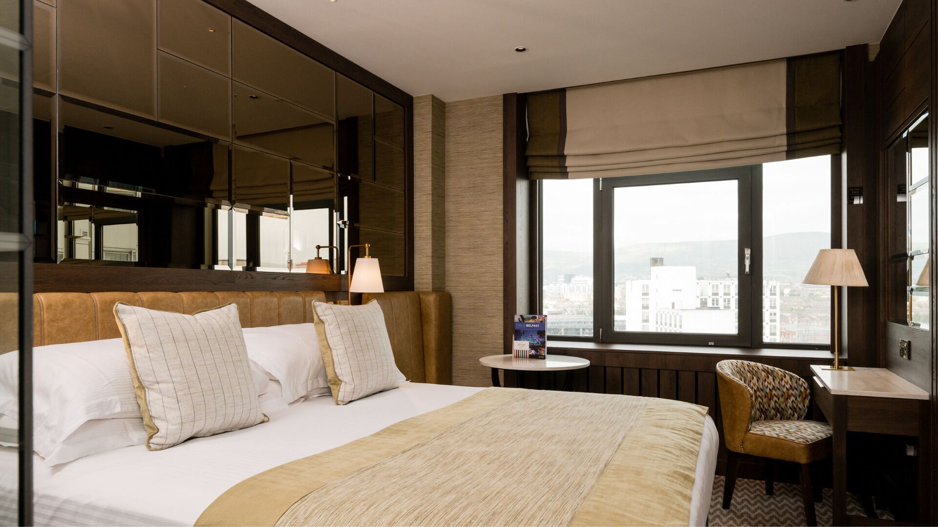 A hotel room with a large bed, a sitting area with a chair, a small table, and a large window offering a city view.