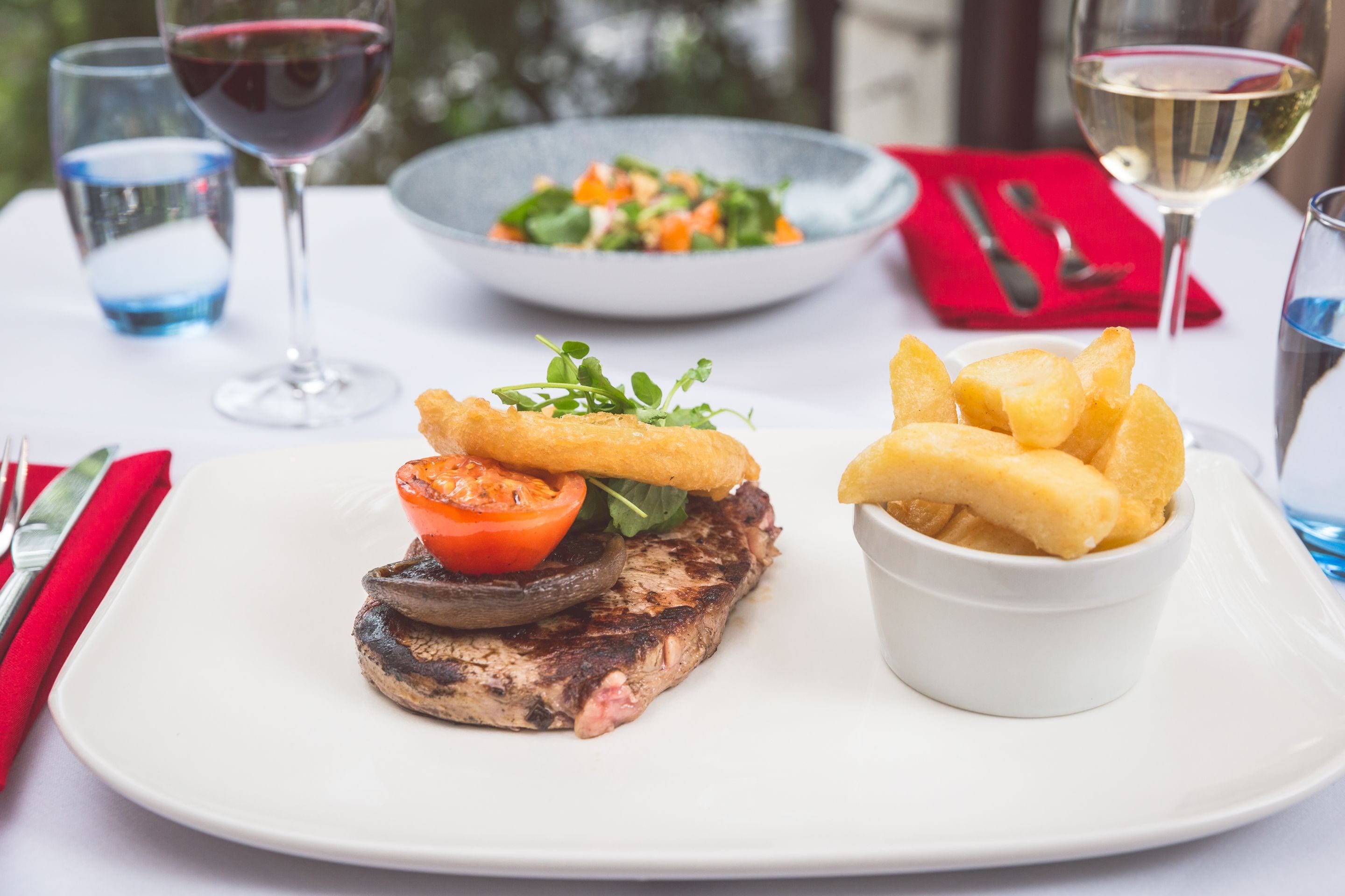 A plate with grilled steak, a tomato, and mushrooms, accompanied by a bowl of fries and a glass of white wine.
