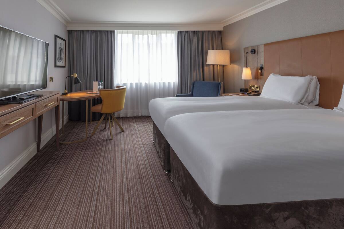 A hotel room with two beds, a desk with a chair, a TV, and a lamp.