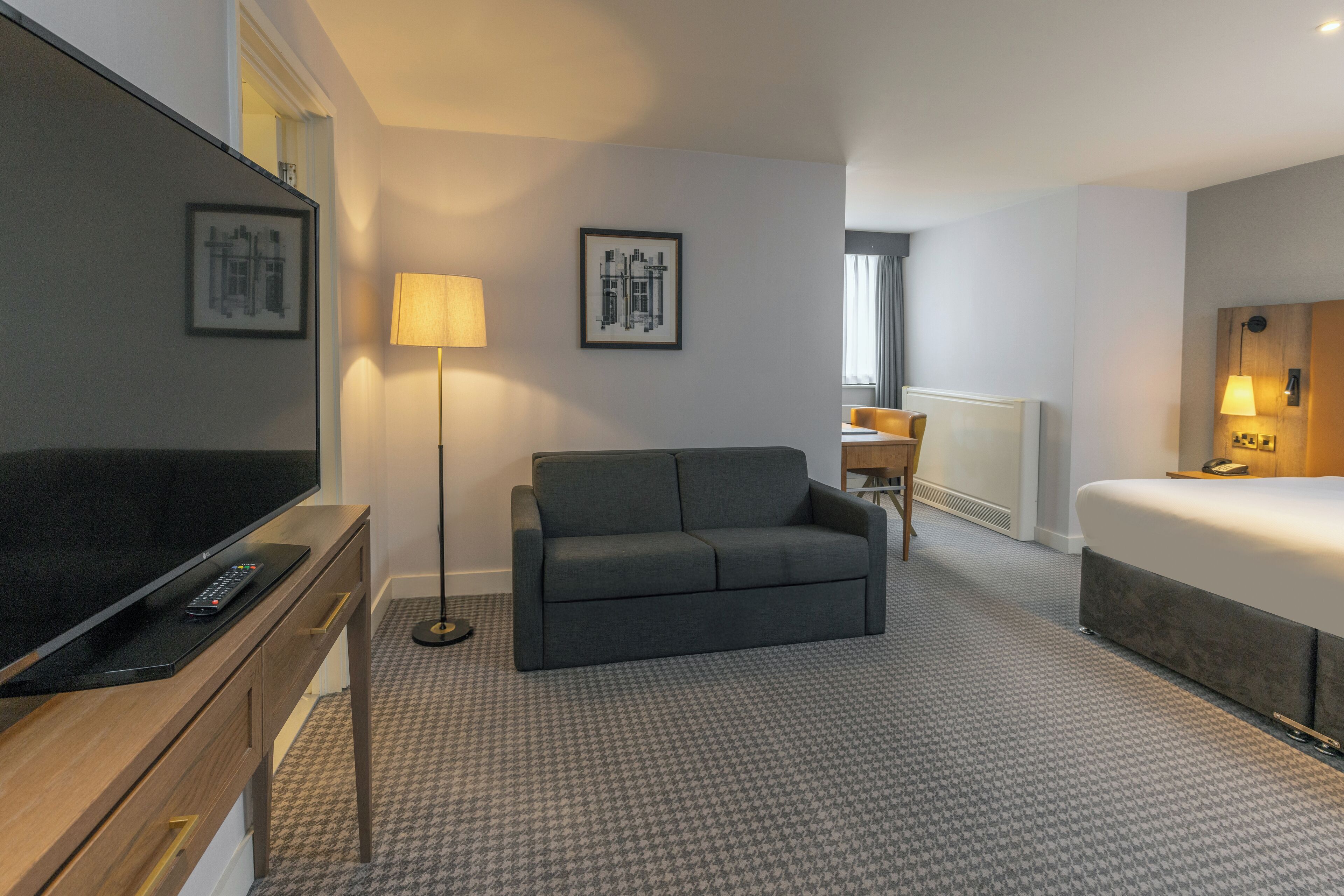 Una camera d'hotel con una TV a schermo piatto, un divano grigio, un letto e due quadri appesi al muro.