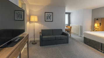 Habitación de hotel con televisión de pantalla plana, un sofá gris, una cama y dos cuadros en la pared.