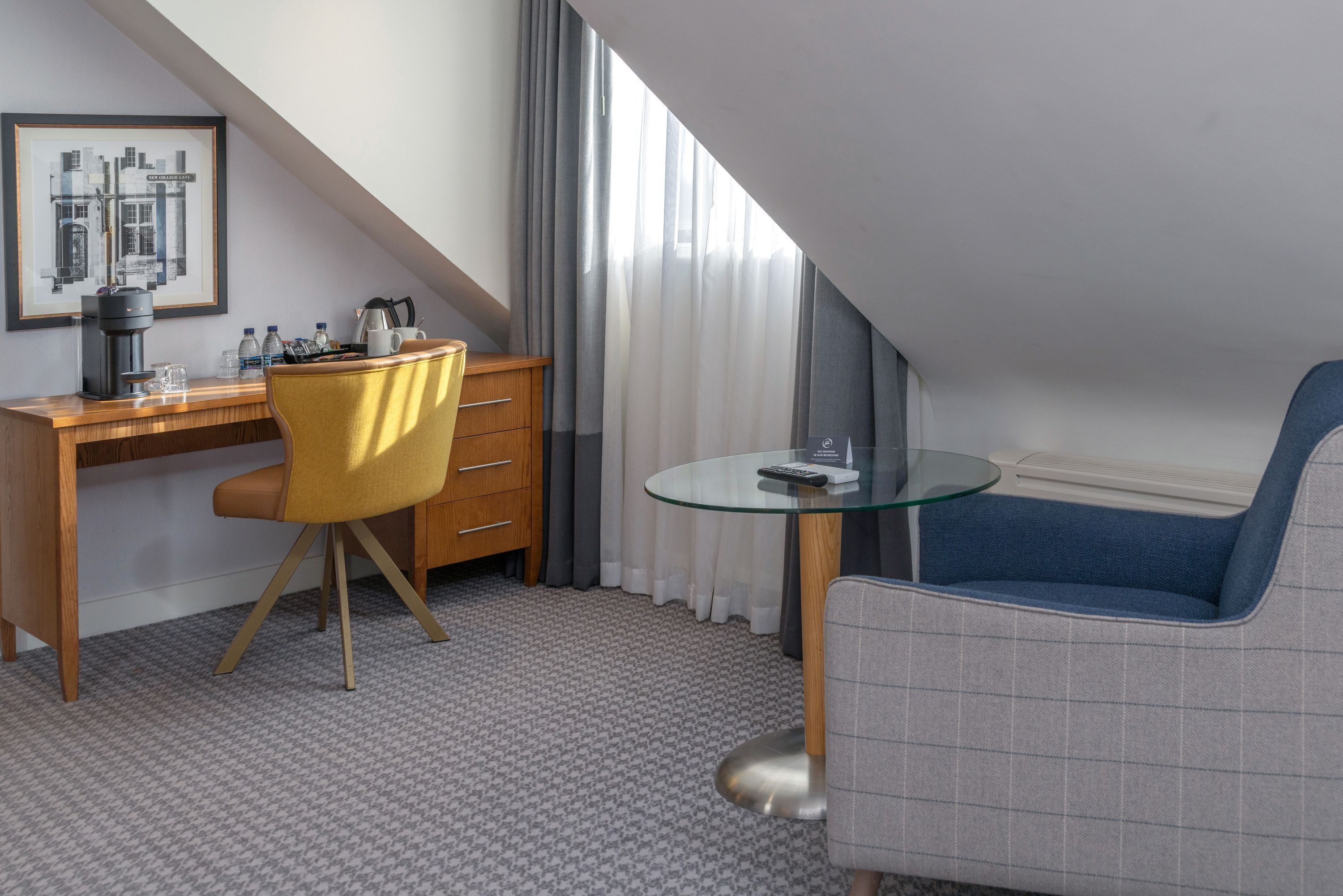 A hotel room with a desk, a chair, a sofa, a glass table, a coffee maker, and a framed cityscape picture.
