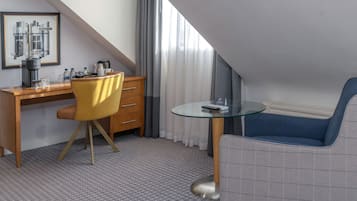 A hotel room with a desk, a chair, a sofa, a glass table, a coffee maker, and a framed cityscape picture.