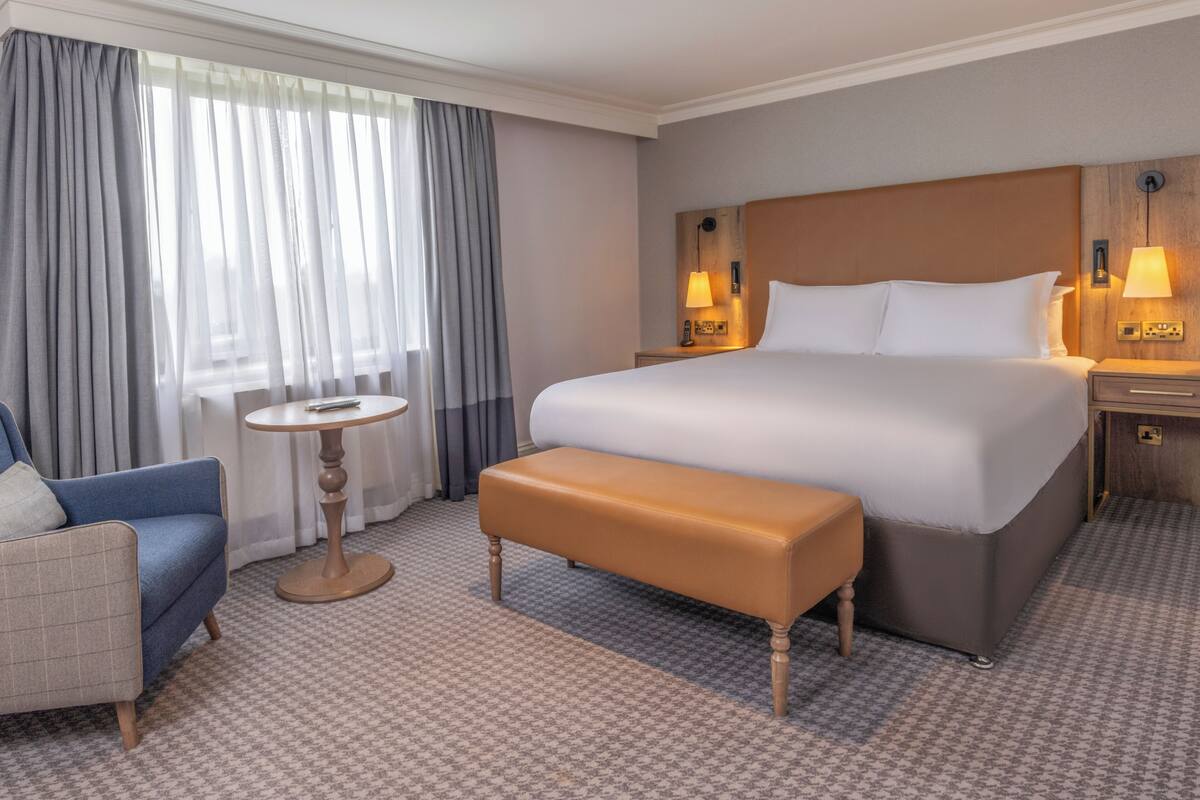 A hotel room with a large bed, a bench, a small table, a chair, and a lamp on the nightstand.