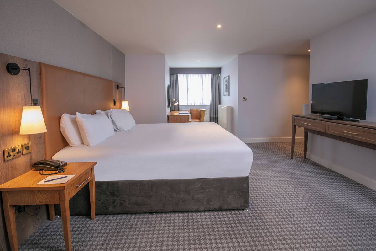 A hotel room with a large bed, bedside lamps, a desk with a telephone, a television, and a view of the room through a window.