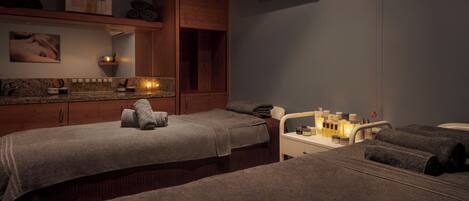 A spa room with two massage tables, towels, a mirror, a shelf with bottles, and a wall-mounted air conditioner.