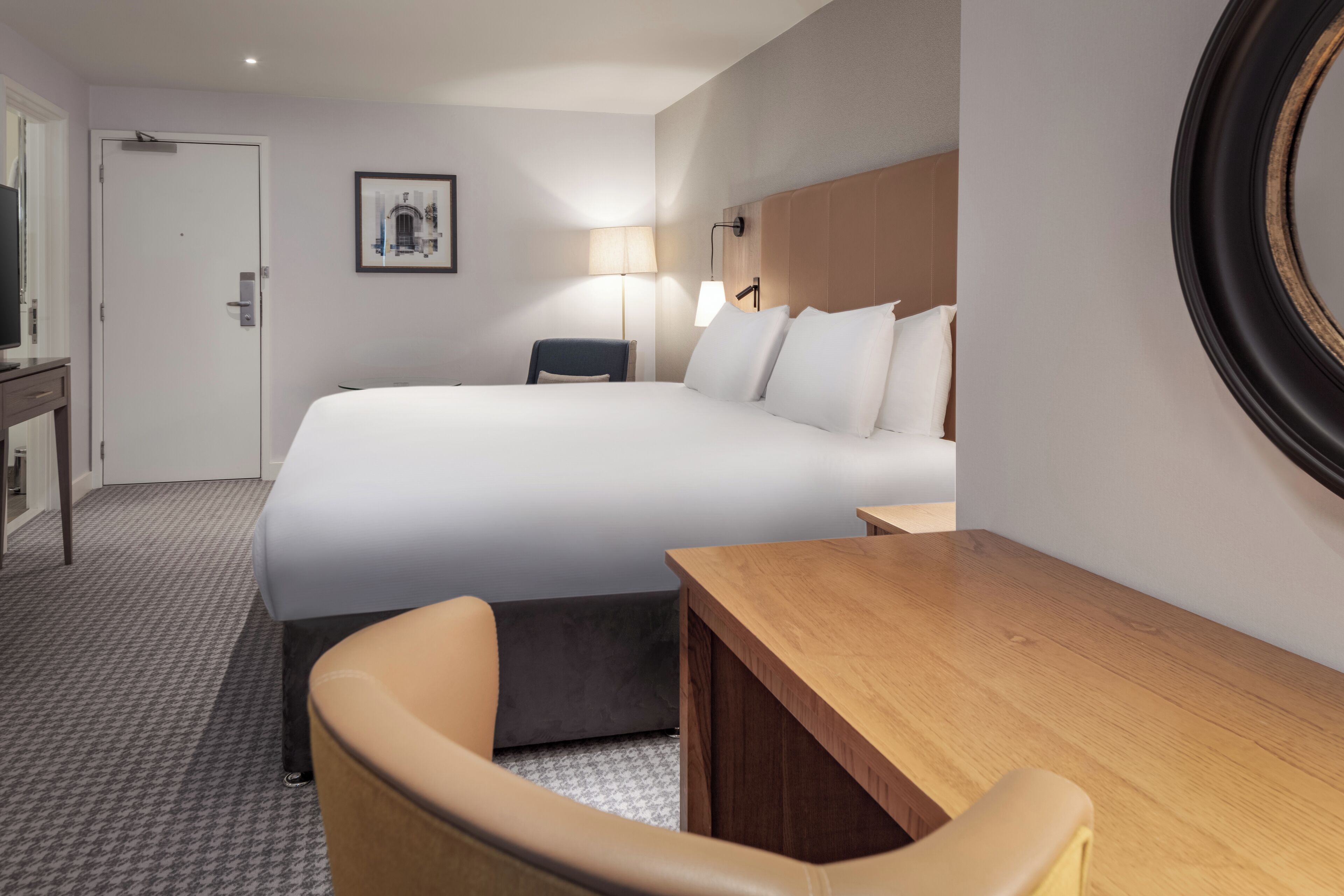 A hotel room with a large bed, a desk, a chair, a lamp, and a framed picture on the wall.