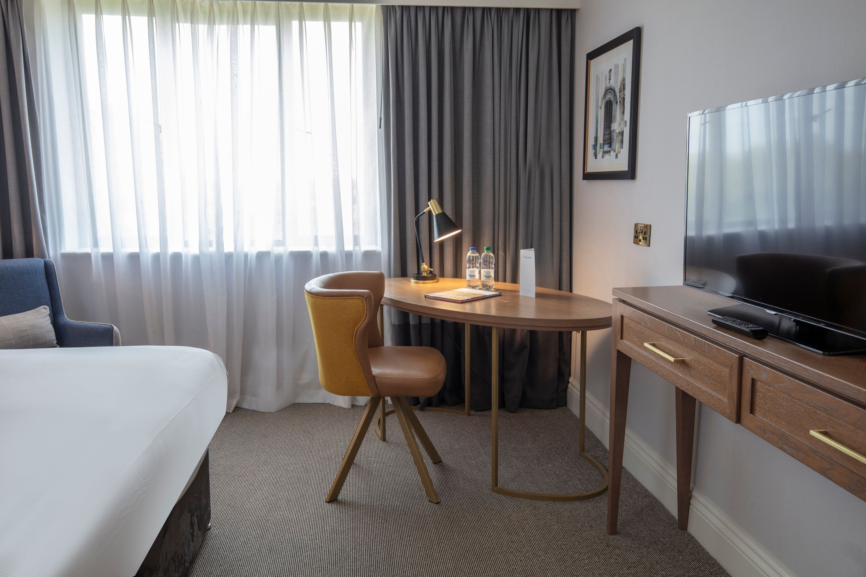 Una habitación de hotel con una cama, un escritorio con lámpara, una silla, un televisor y una ventana con cortinas.