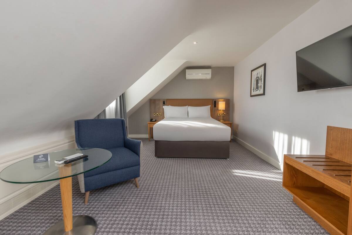 A hotel room with a bed, a blue armchair, a glass table, a wooden bench, and a flat-screen TV on the wall.