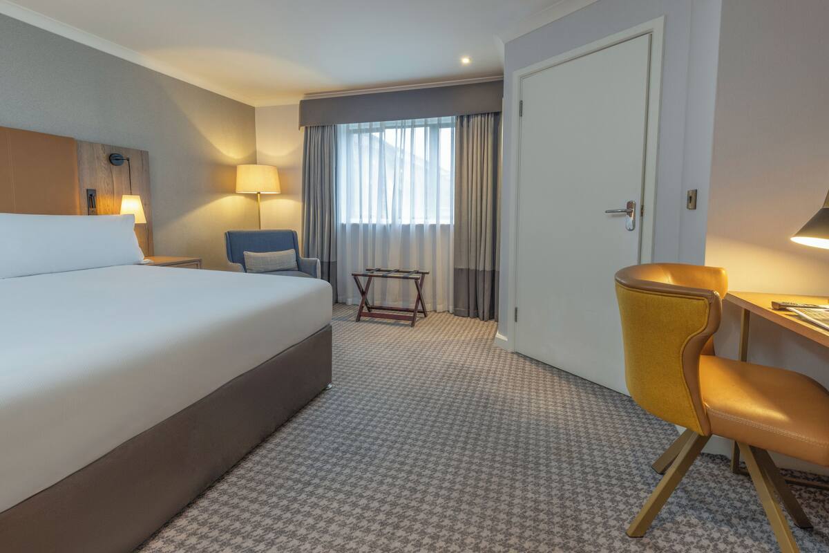 A hotel room with a large bed, a desk with a chair, a small table, and a window with curtains.
