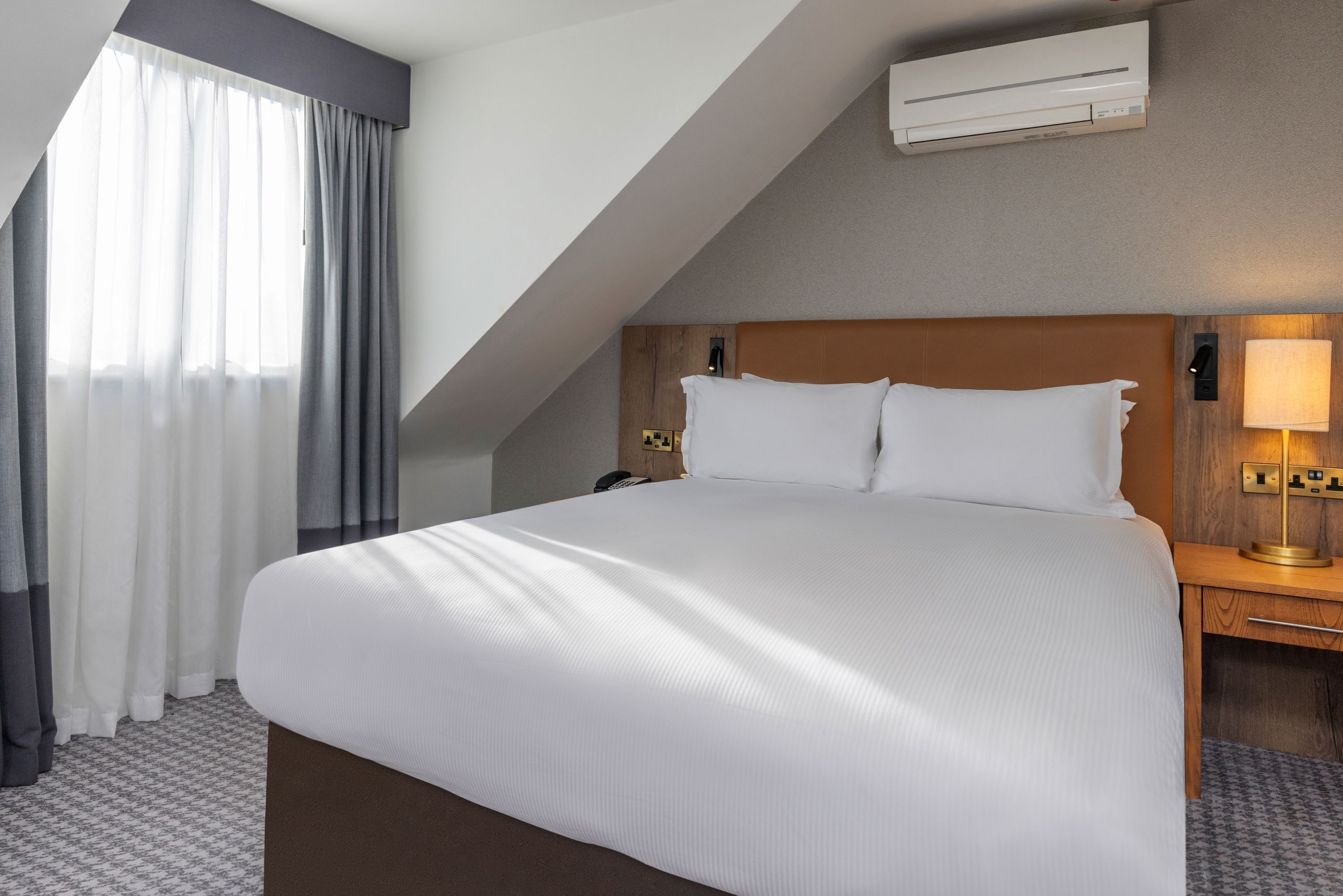 A hotel room with a large bed, a wooden headboard, a nightstand with a lamp, and a window with curtains.