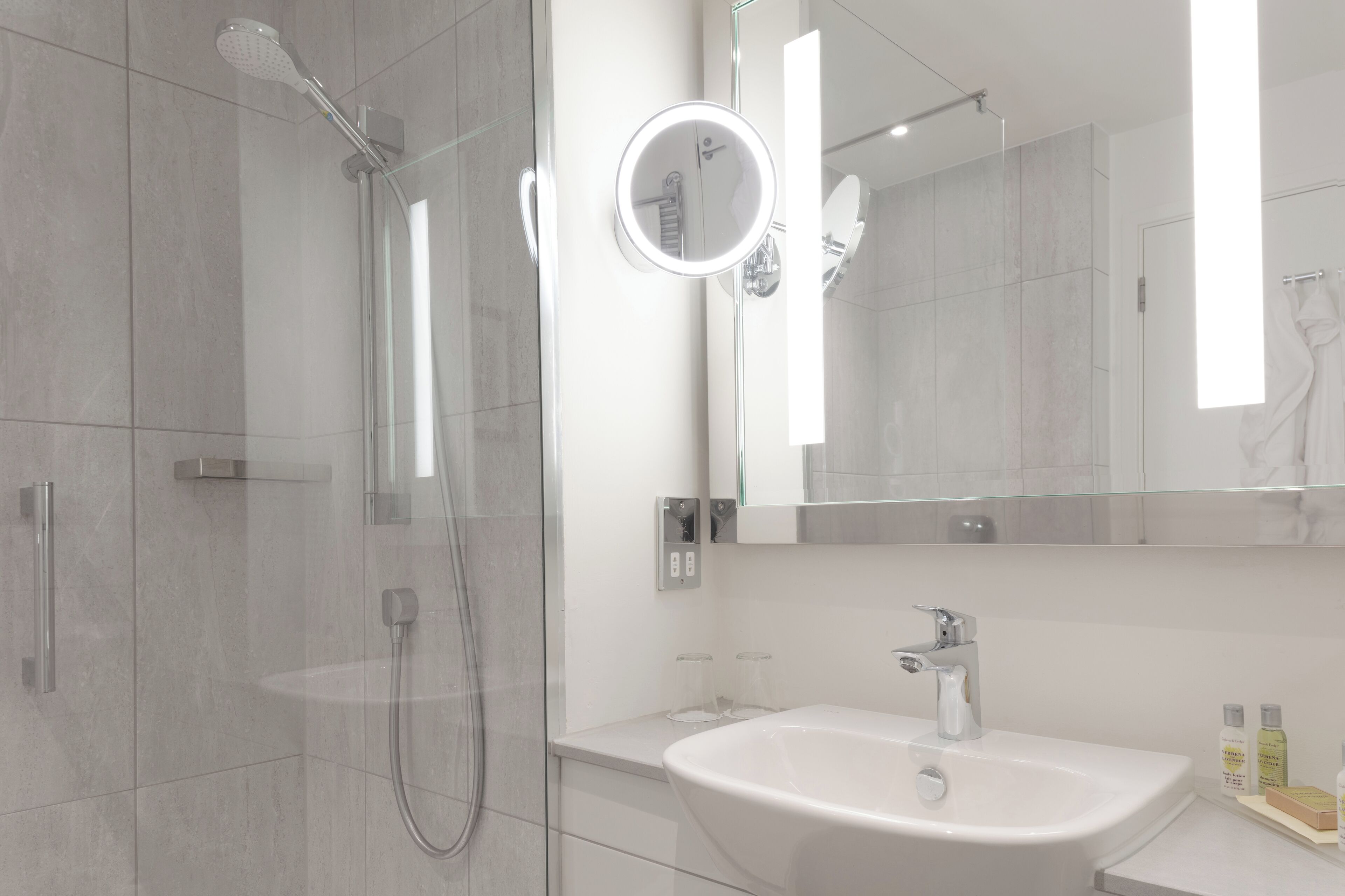 A modern bathroom with a glass-enclosed shower, a white sink, and a large mirror with a round light.