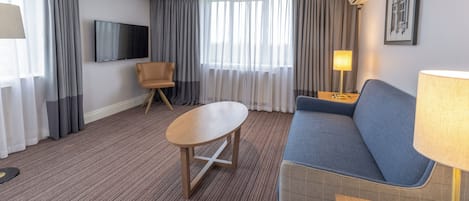 A hotel room with a blue sofa, a wooden coffee table, a TV mounted on the wall, and a large window with curtains.