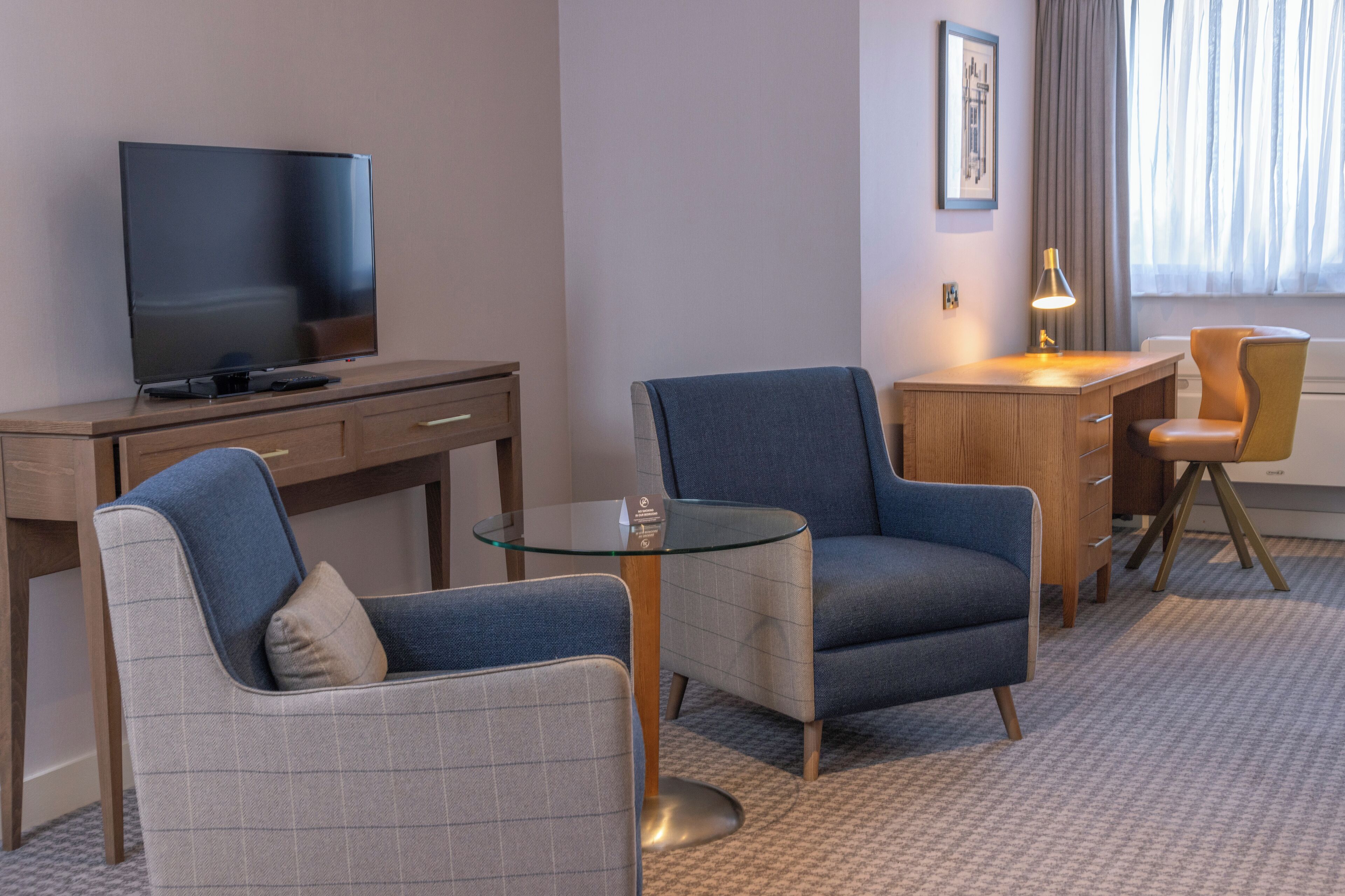 A hotel room with a flat-screen TV, a wooden desk, two armchairs, a glass coffee table, and a lamp.