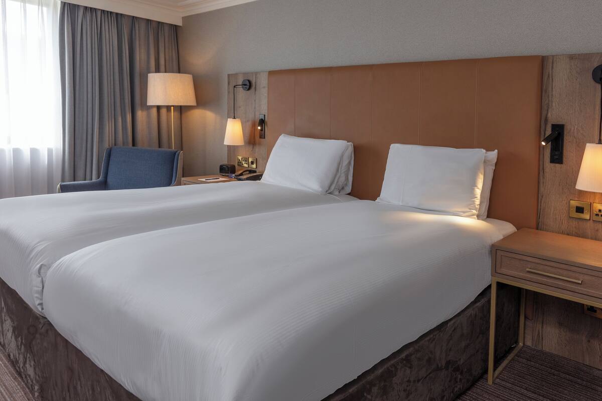 A double bed with white linens, a wooden headboard, bedside lamps, and a chair.