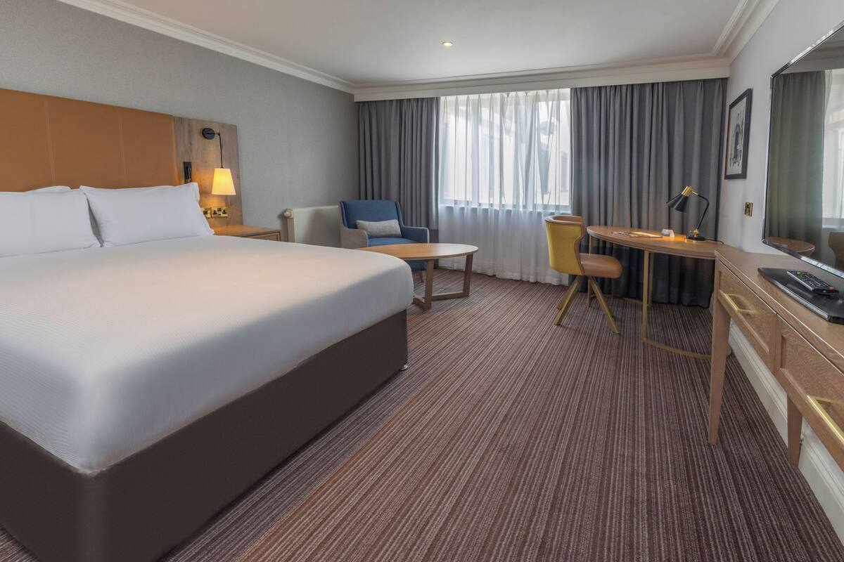 A hotel room with a large bed, a desk with a chair, a small table, and a TV.