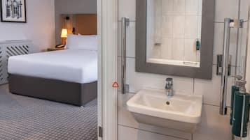 Habitación de hotel con cama, mesita de noche, lámpara y baño con lavamanos y espejo.