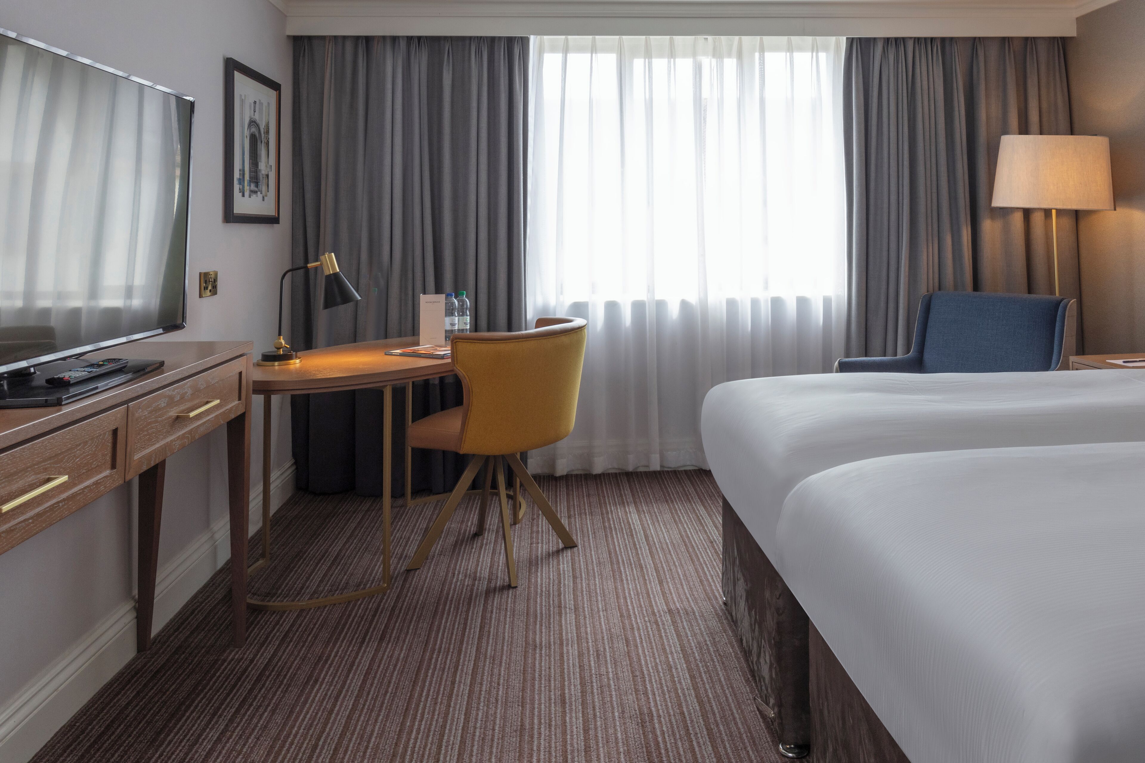 A hotel room with a bed, a desk with a chair, a television, a lamp, and a window with curtains.