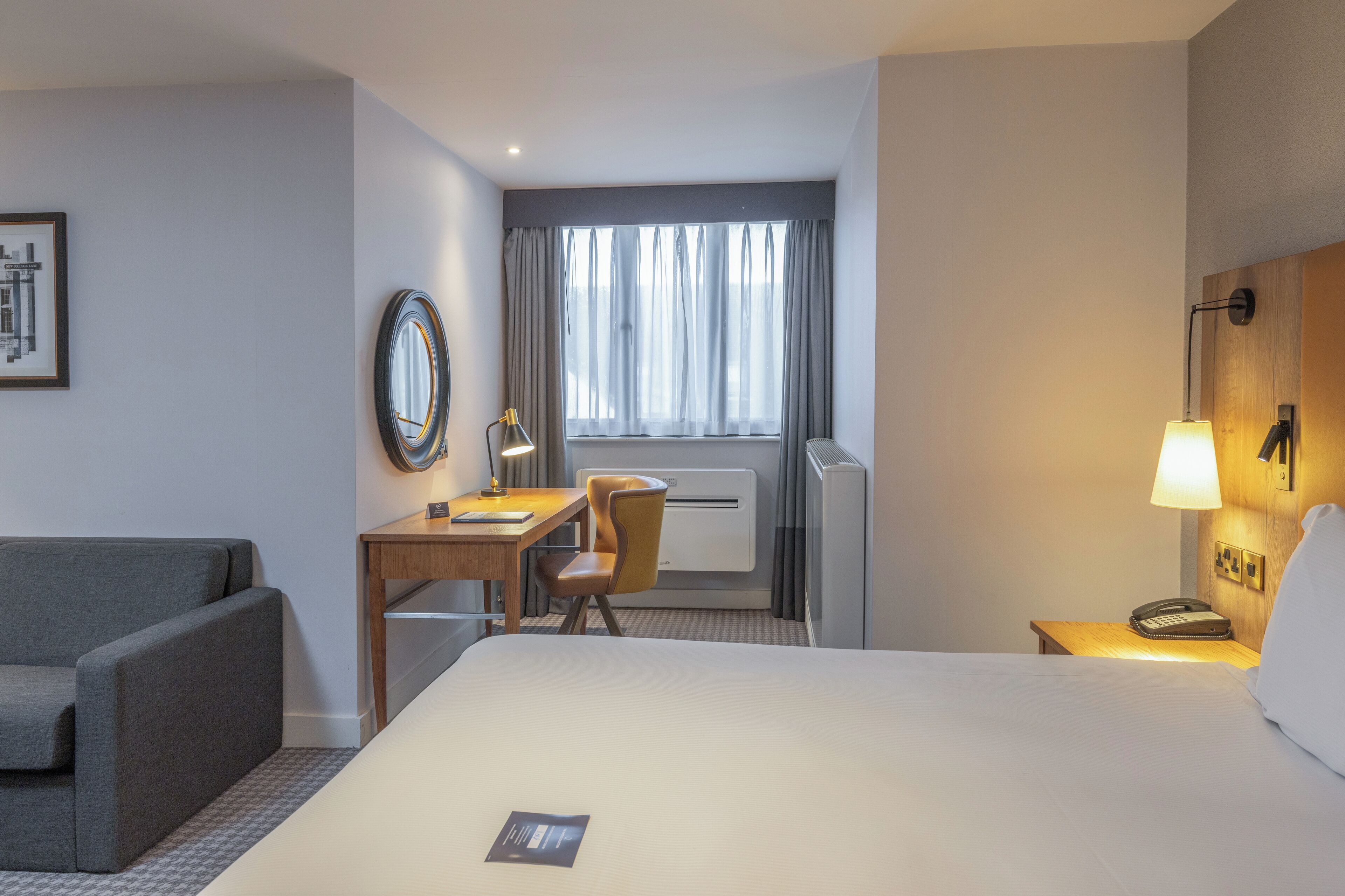 Habitación de hotel con cama, escritorio, silla, sofá, lámpara, teléfono y ventana con cortinas.