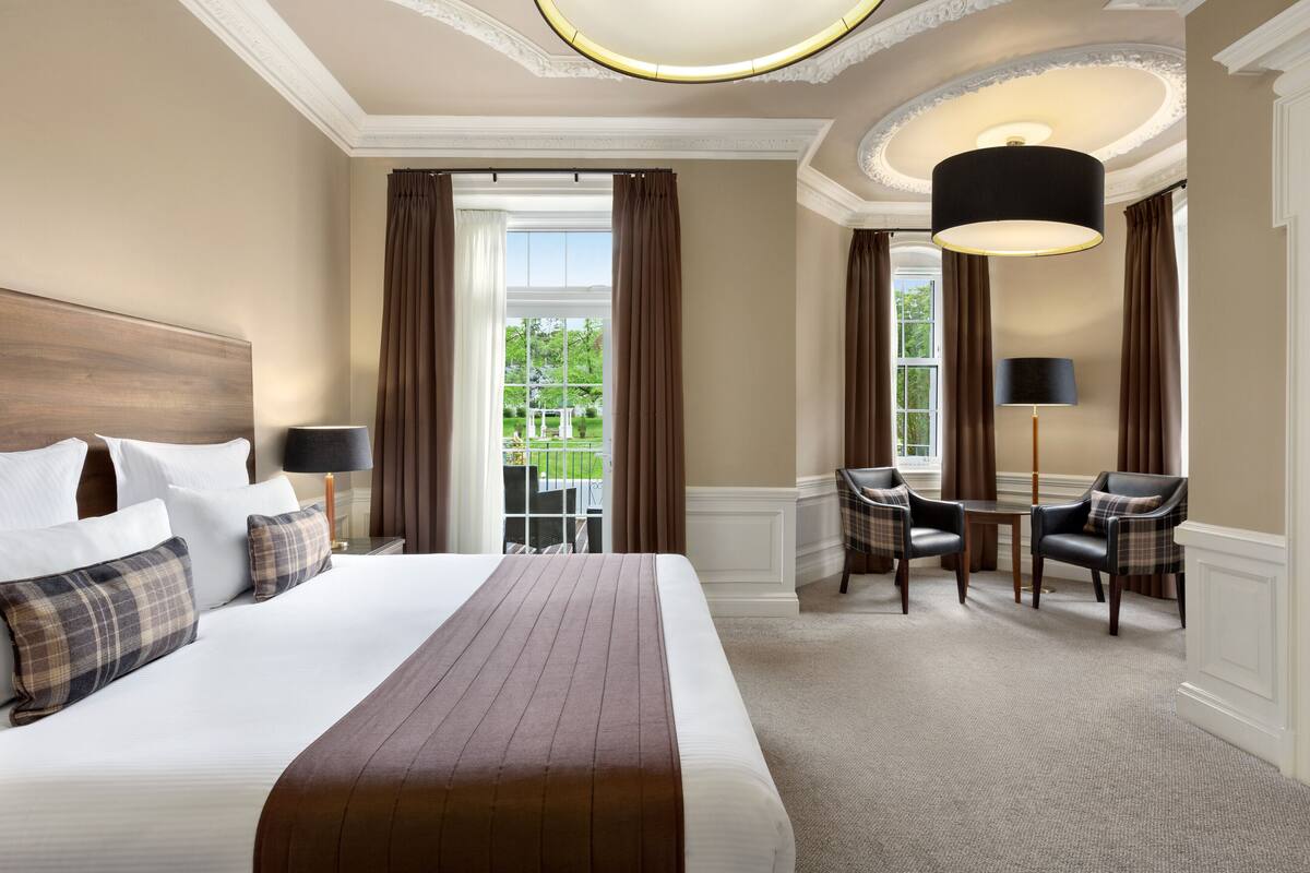 Une chambre d’hôtel avec un grand lit, un bureau, deux chaises et une vue sur la verdure.