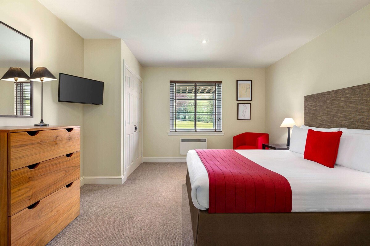Een hotelkamer met een bed, een dressoir, een televisie, een raam met zonwering en een rode stoel.