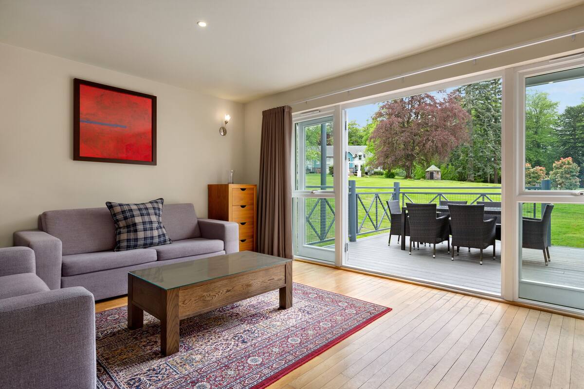Un salon moderne avec un canapé, une table basse en verre et une vue sur une terrasse extérieure aménagée avec des chaises.