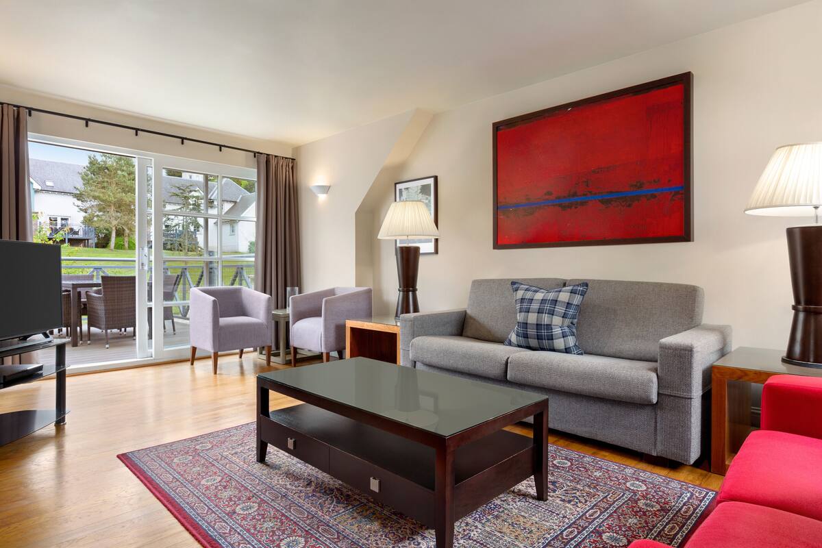 Un salon moderne avec un canapé gris, une table basse en verre et un fauteuil rouge. On y trouve une grande fenêtre avec des rideaux, un téléviseur à écran plat et un tableau abstrait rouge accroché au mur.