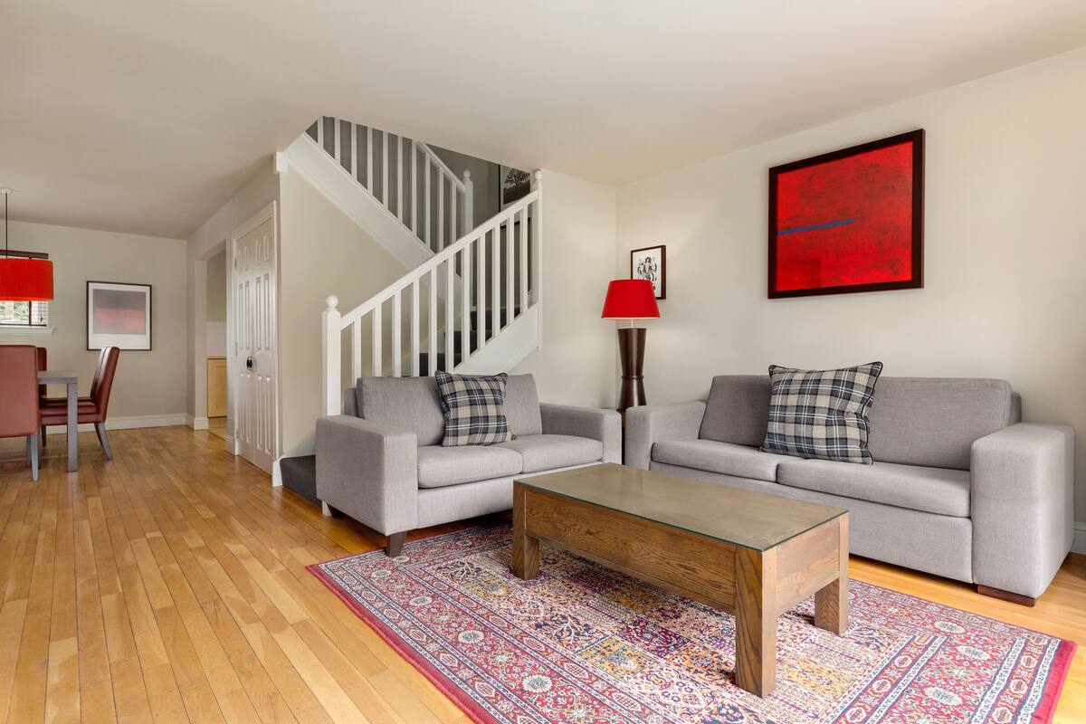 Un salon avec deux canapés gris, une table basse en bois, un tapis aux motifs rouges et bleus, un escalier avec des rampes blanches et un tableau abstrait rouge accroché au mur.