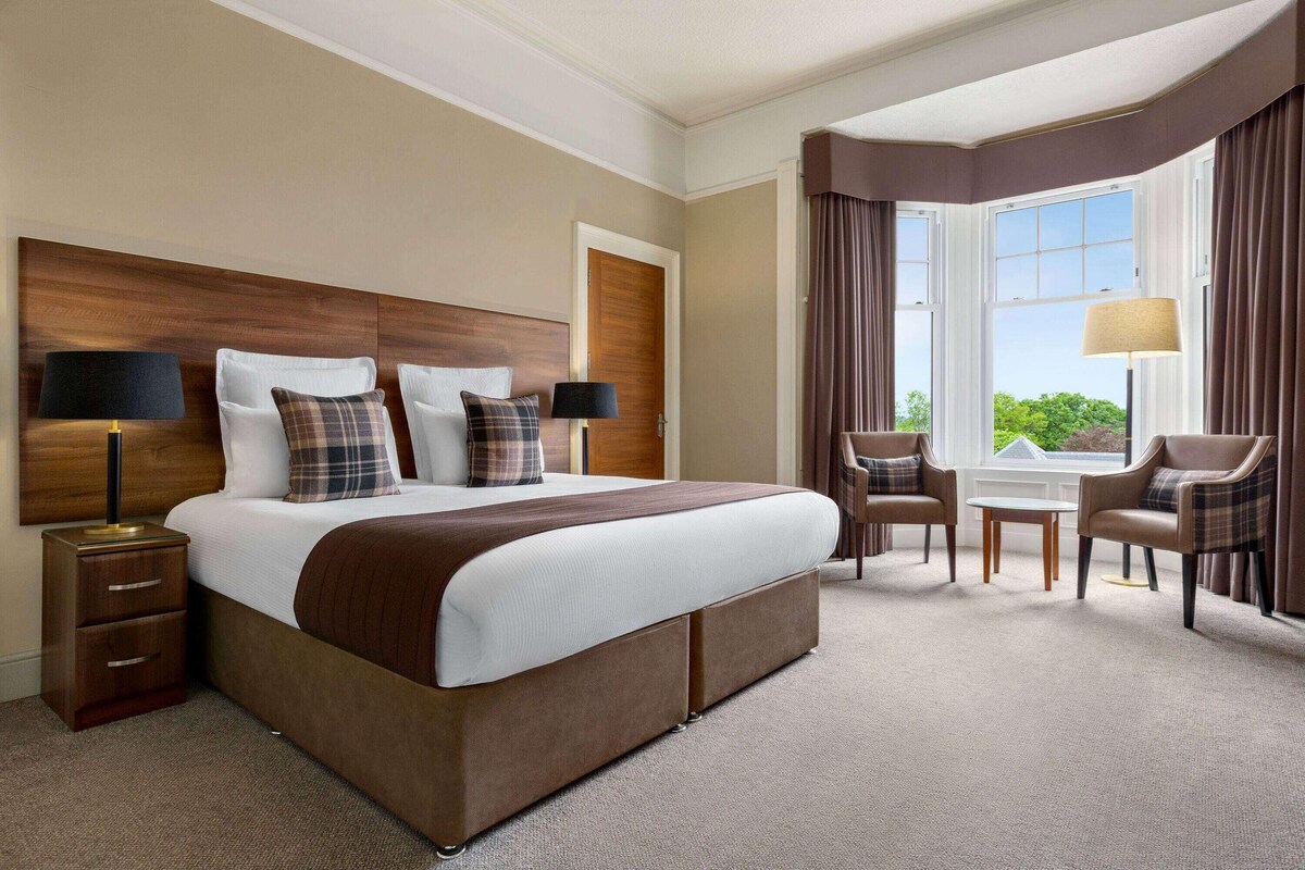 Een hotelkamer met een groot bed, twee nachtkastjes, een bureau, twee stoelen en een raam met gordijnen.