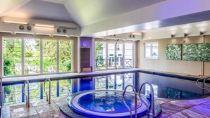 Une grande piscine intérieure avec un jacuzzi, entourée de grandes fenêtres donnant sur un paysage verdoyant.