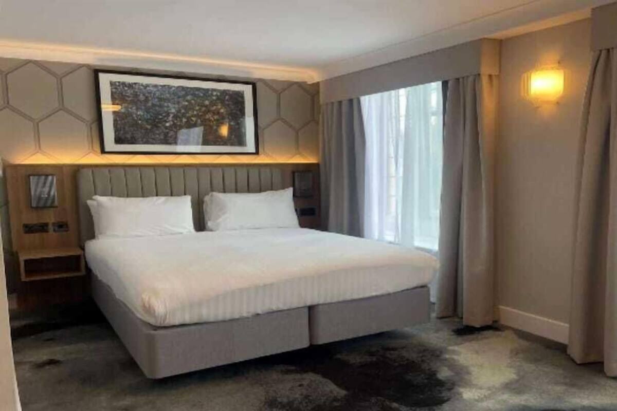 A modern hotel room with a large bed, a headboard with built-in lighting, a framed artwork above the bed, and a large window with curtains.