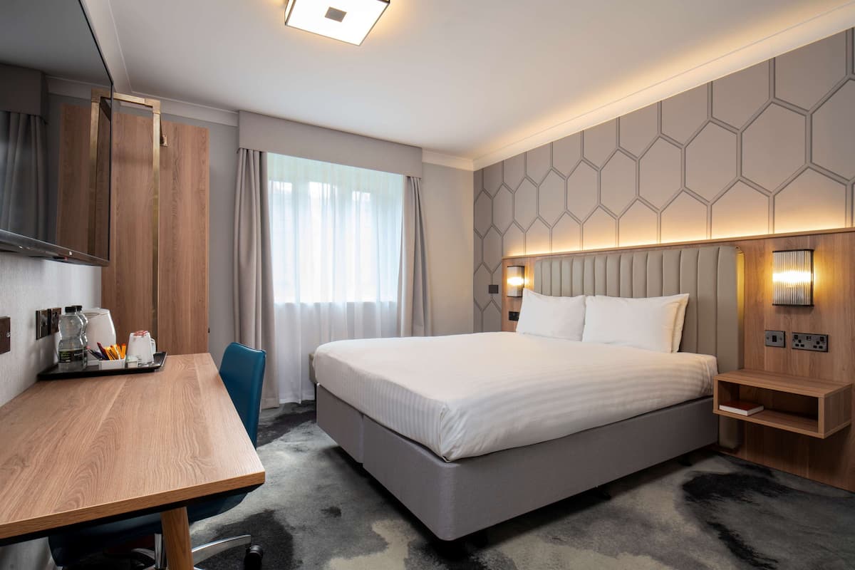A modern hotel room with a large bed, a desk, a chair, a mirror, and a nightstand with a lamp.