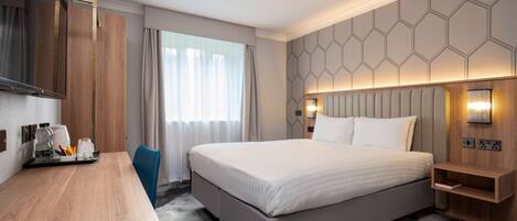 A modern hotel room with a large bed, a desk, a chair, a mirror, and a nightstand with a lamp.