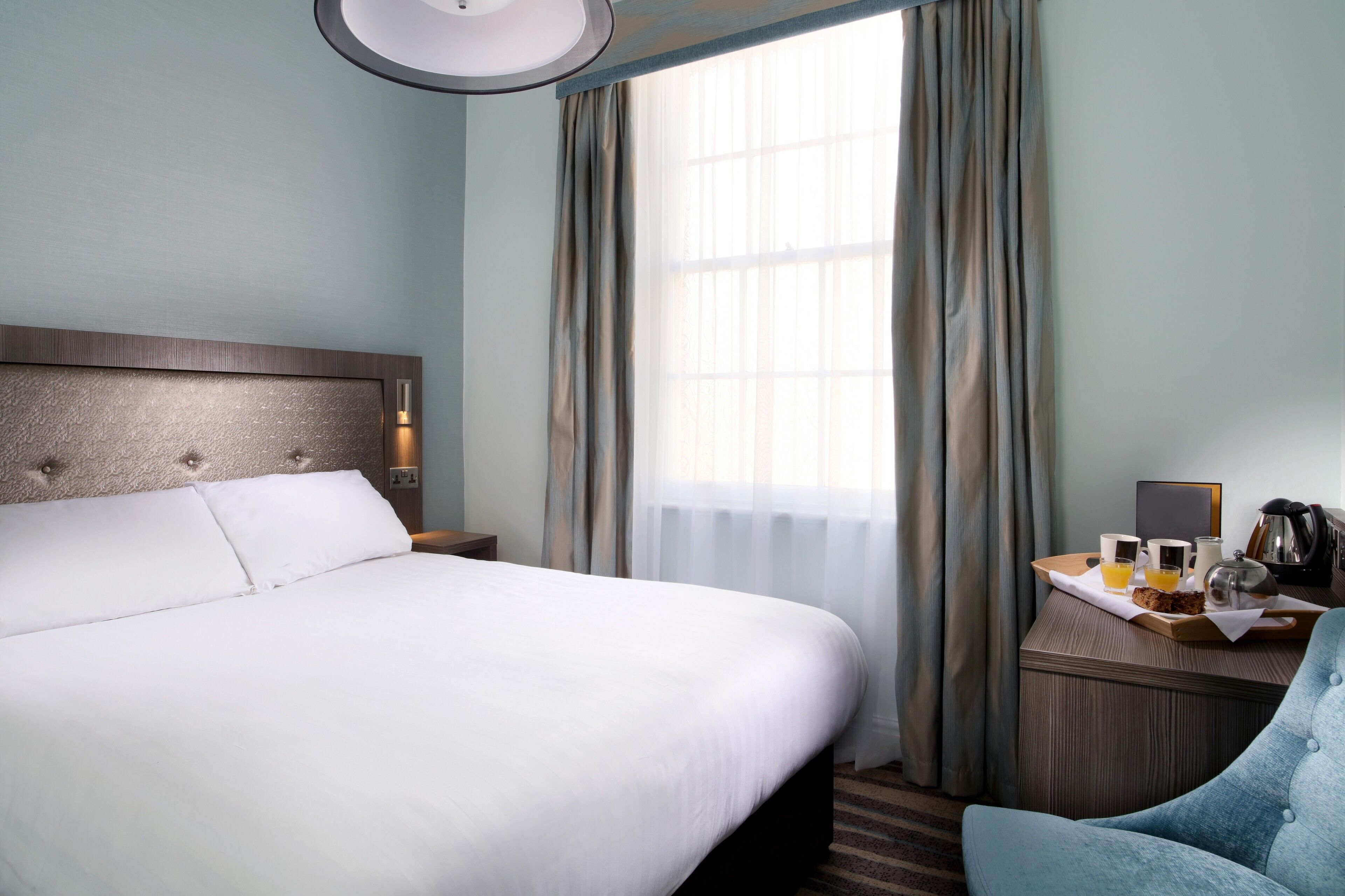 Une chambre d’hôtel avec un grand lit, une table de chevet avec une lampe, une chaise et un plateau de petit-déjeuner avec une machine à café.