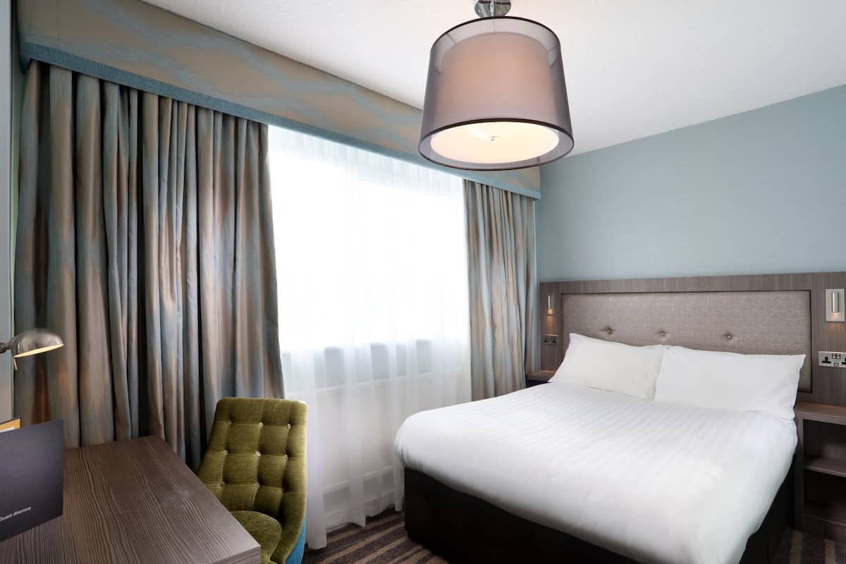 A hotel room with a large bed, a desk, a chair, and a lamp.