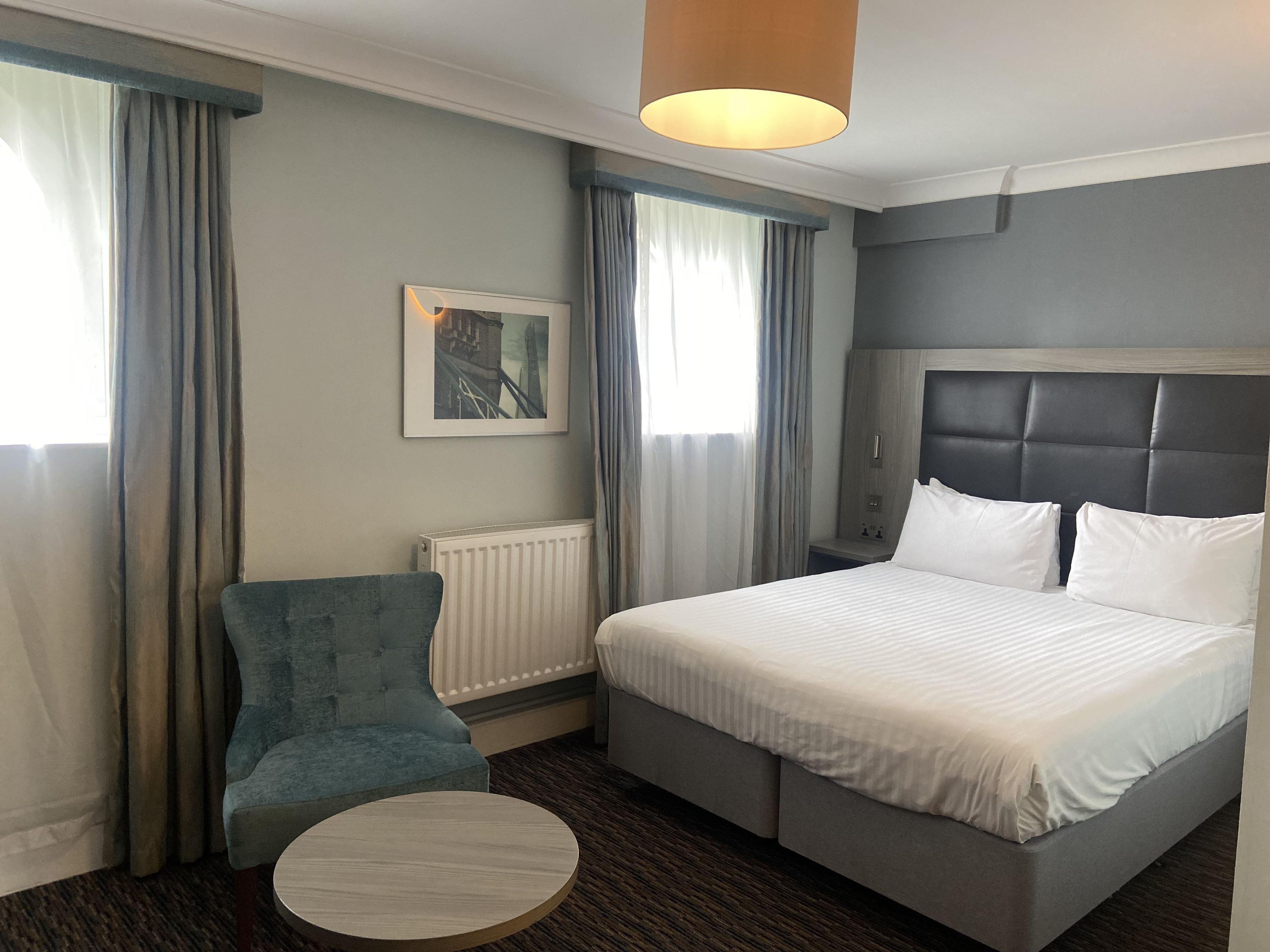 Una camera d'albergo con un letto, un tavolino rotondo, una poltrona blu, un termosifone e un quadro appeso al muro.