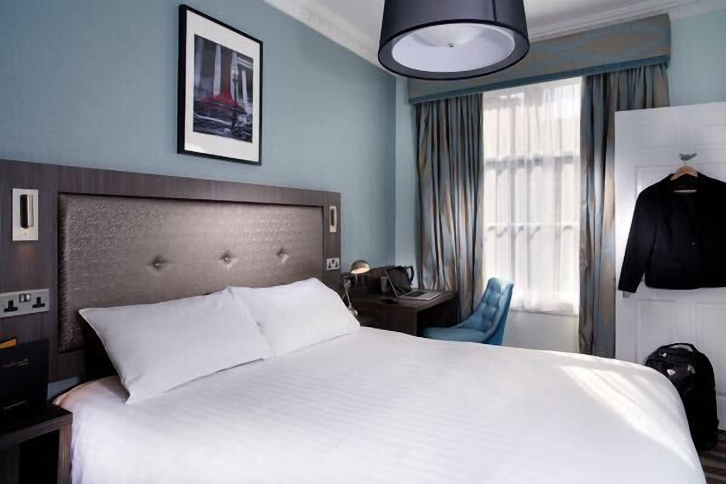 A hotel room with a large bed, a desk with a lamp, a chair, a coat rack, and a window with curtains.