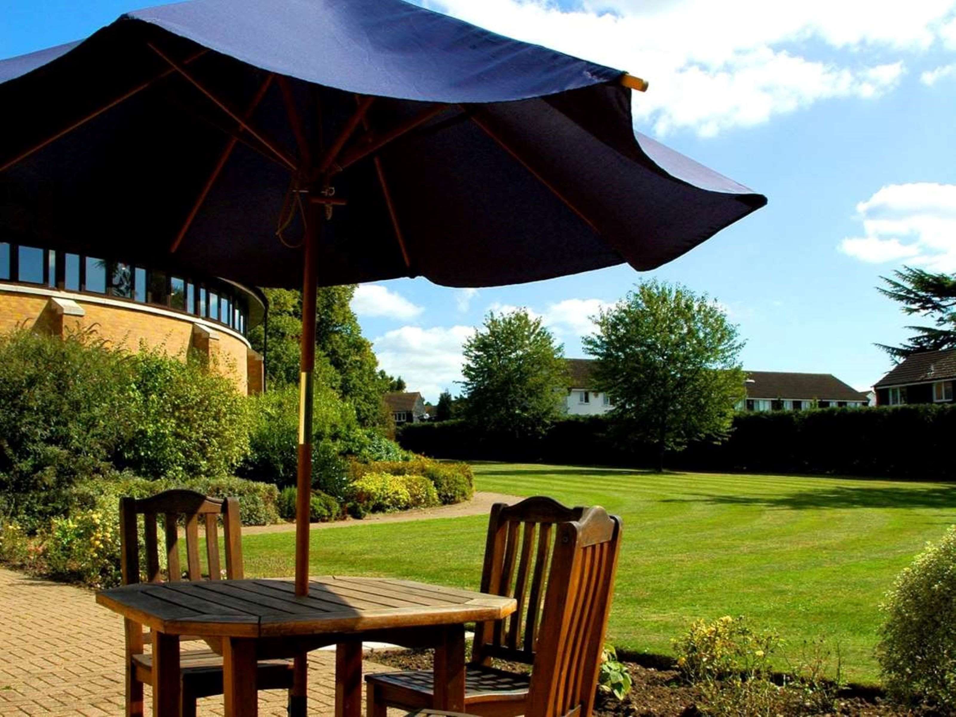 An outdoor seating area with a table and chairs under a large umbrella, surrounded by a well-maintained lawn and a building in the background.