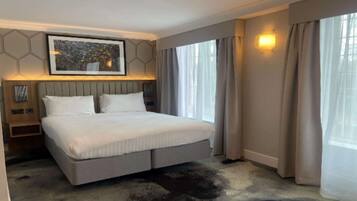 A modern hotel room with a large bed, a headboard with built-in lighting, a framed artwork above the bed, and a large window with curtains.