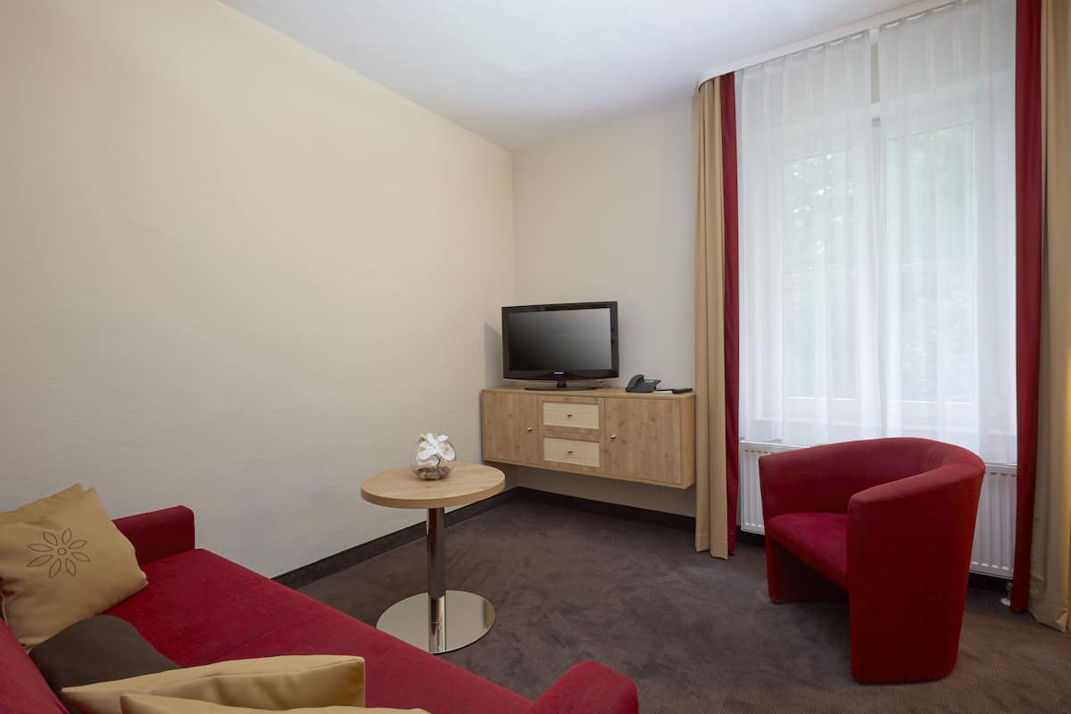 A hotel room with a red sofa, a small round table, a television on a wooden cabinet, and a large window with curtains.