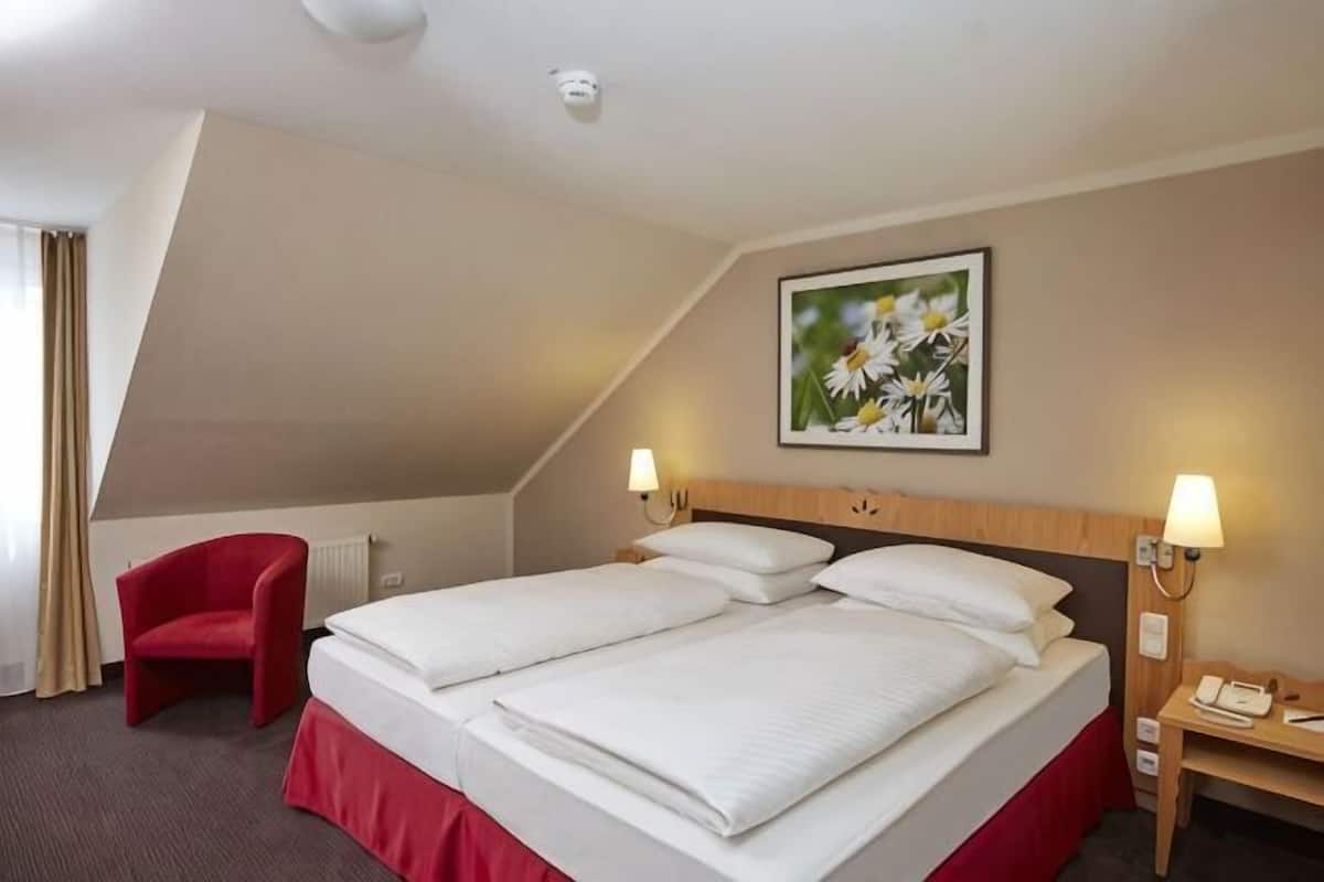 A hotel room with a bed, a red chair, a desk, a lamp, and a painting on the wall.