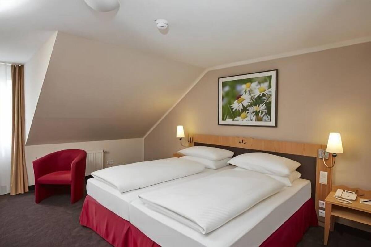 A hotel room with a bed, a red chair, a table with a lamp, and a painting on the wall.