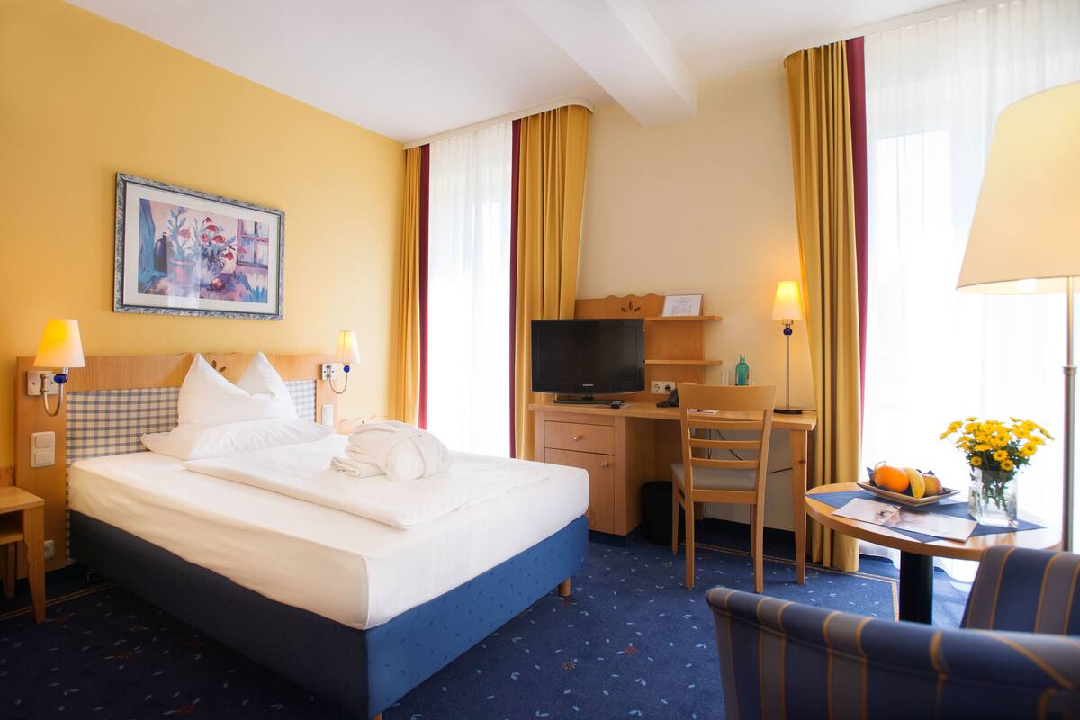 A hotel room with a bed, desk, chair, TV, lamp, and a painting on the wall.