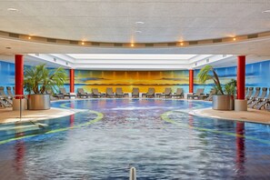 Una gran piscina interior con un mural de atardecer en las paredes, pilares rojos y sillones de descanso alrededor.