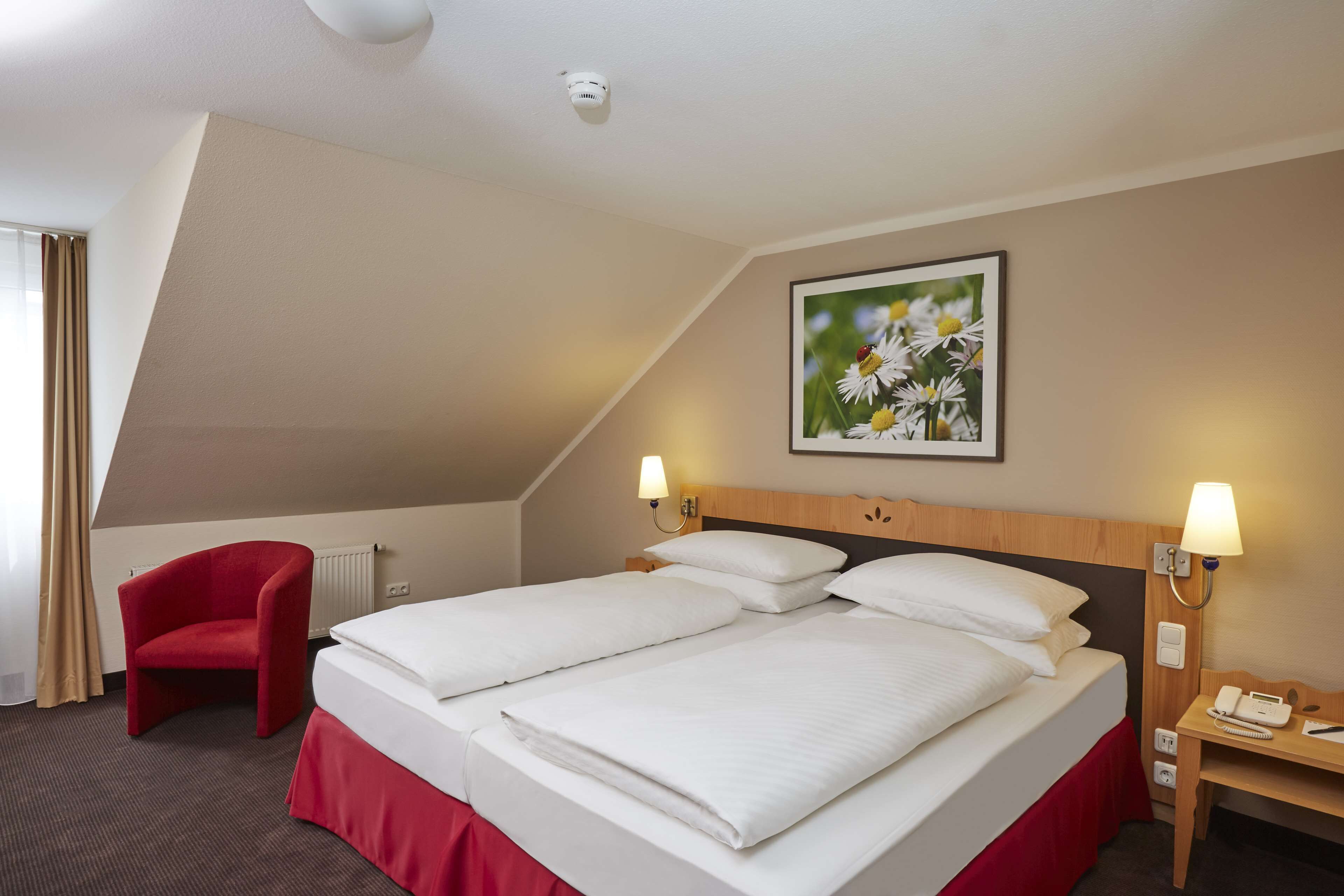 A hotel room with two beds, a red armchair, a nightstand with a lamp, and a framed picture on the wall.